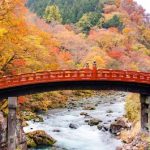 The iconic red painted Shinkyo Bridge with autumn foliage in the hills and a flowing river.