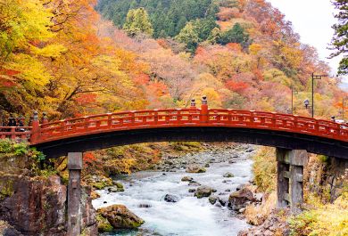 The iconic red painted Shinkyo Bridge with autumn foliage in the hills and a flowing river.