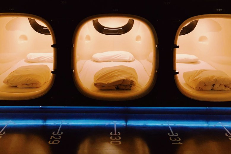 An image of 3 capsule hotel beds illuminated in a dark room.
