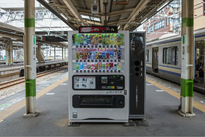 A white Japanese vending machine at Chiba Station, with a train on the right side.